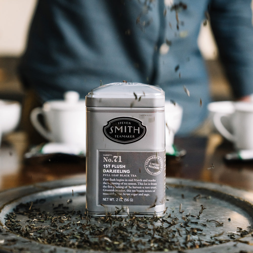 Tin of First Flush Darjeeling on a silver tray with tea leaves falling from above.