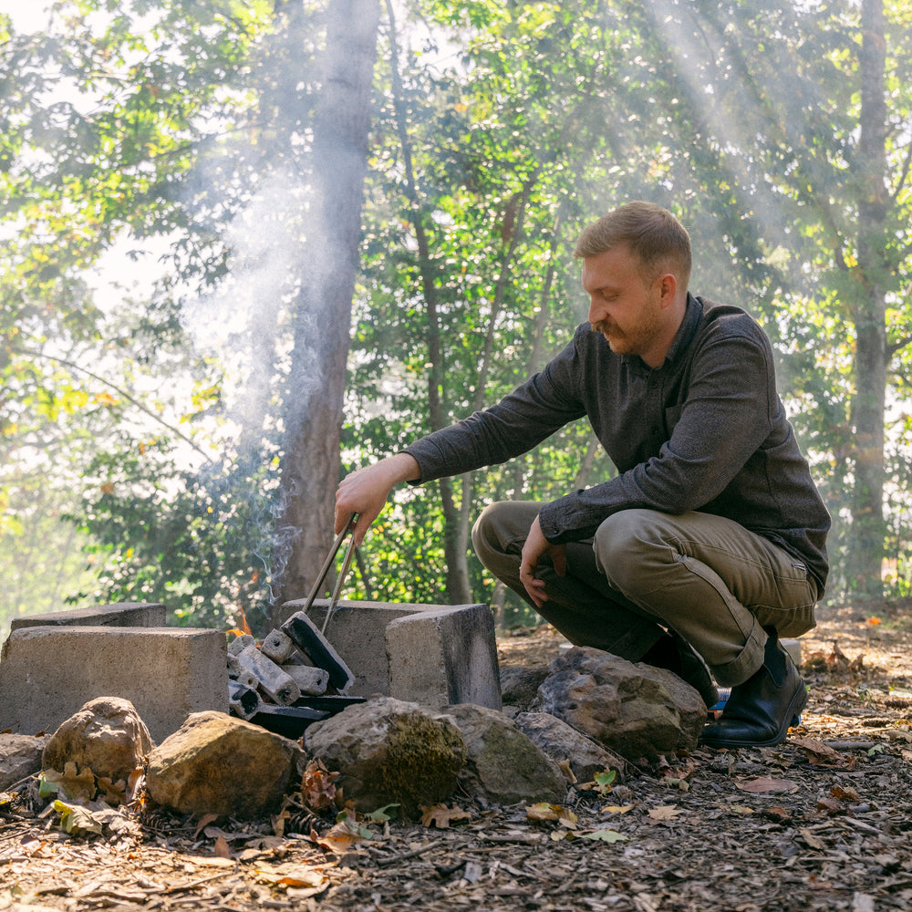 Teamaker crouching beside a charcoal fire in the woods arranging charcoal to stoke fire.
