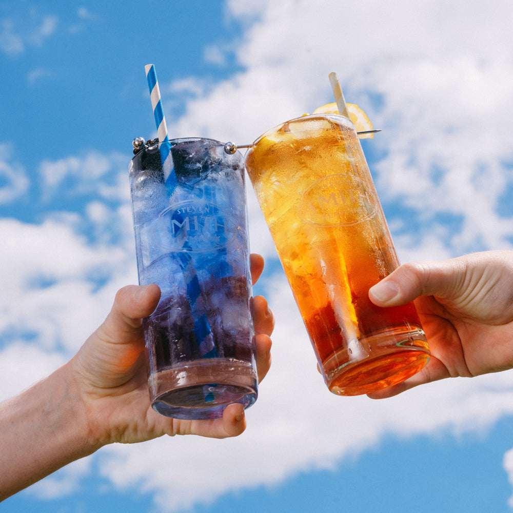 Two hands holding glasses of Blackberry Jasmine Iced Tea and Lemon Black Iced Tea against a blue sky with clouds
