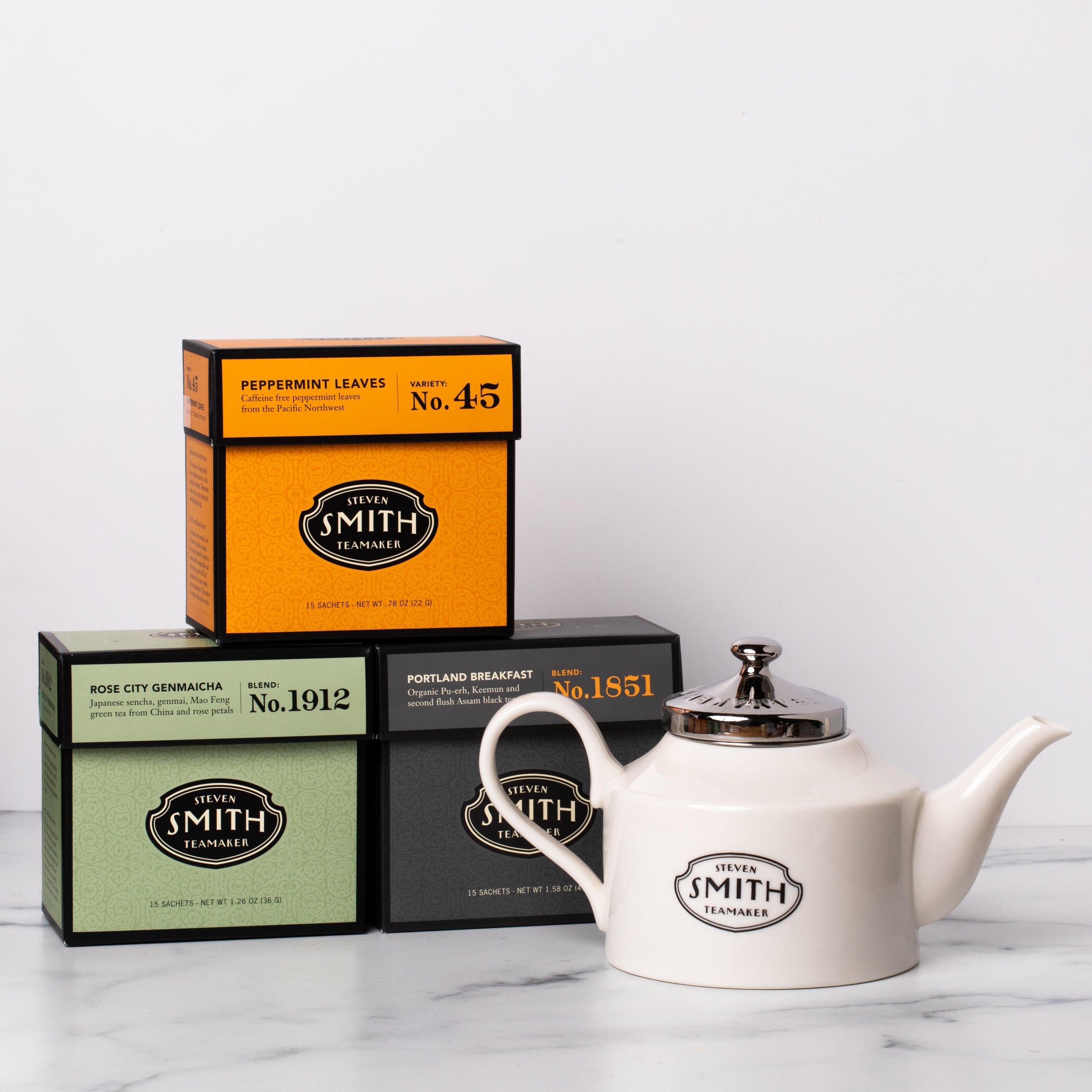 One black carton, one orange carton and one green carton of tea with a white porcelain teapot.