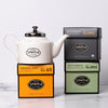 One black carton, one orange carton and one green carton of tea with a white porcelain teapot.