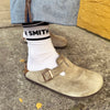 White crew socks with Smith Teamaker written around the ankle in black block letters worn with Birkenstocks and jeans.