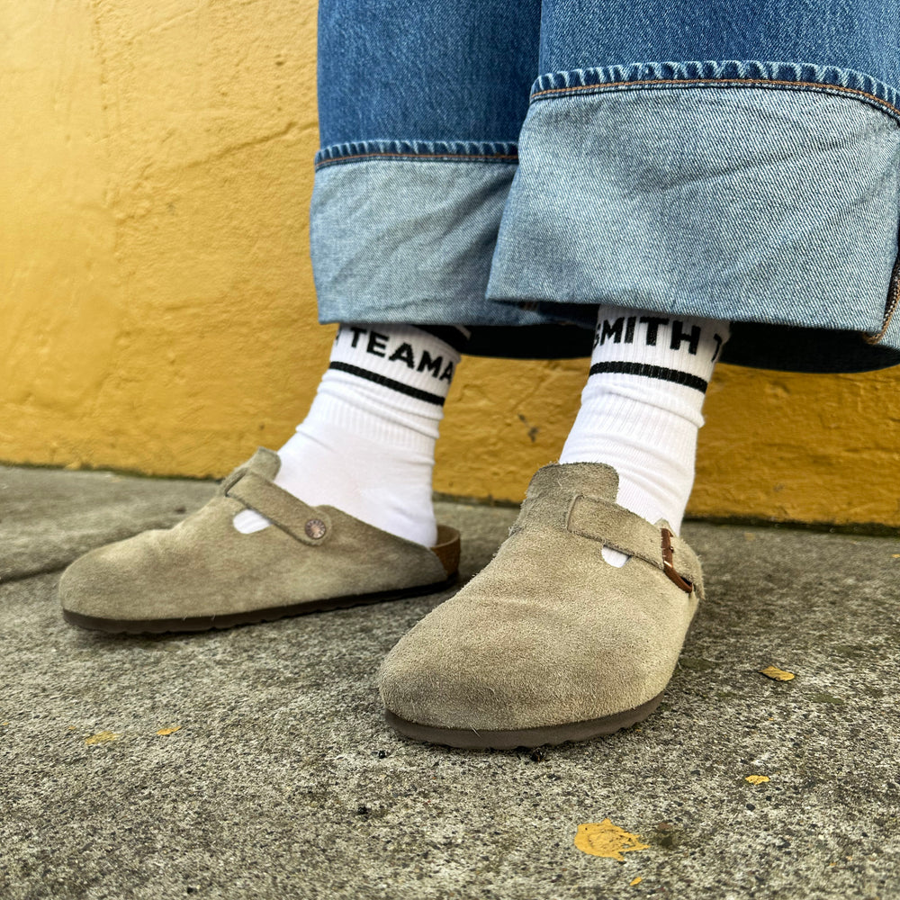 White crew socks with Smith Teamaker written around the ankle in black block letters worn with Birkenstocks and jeans.