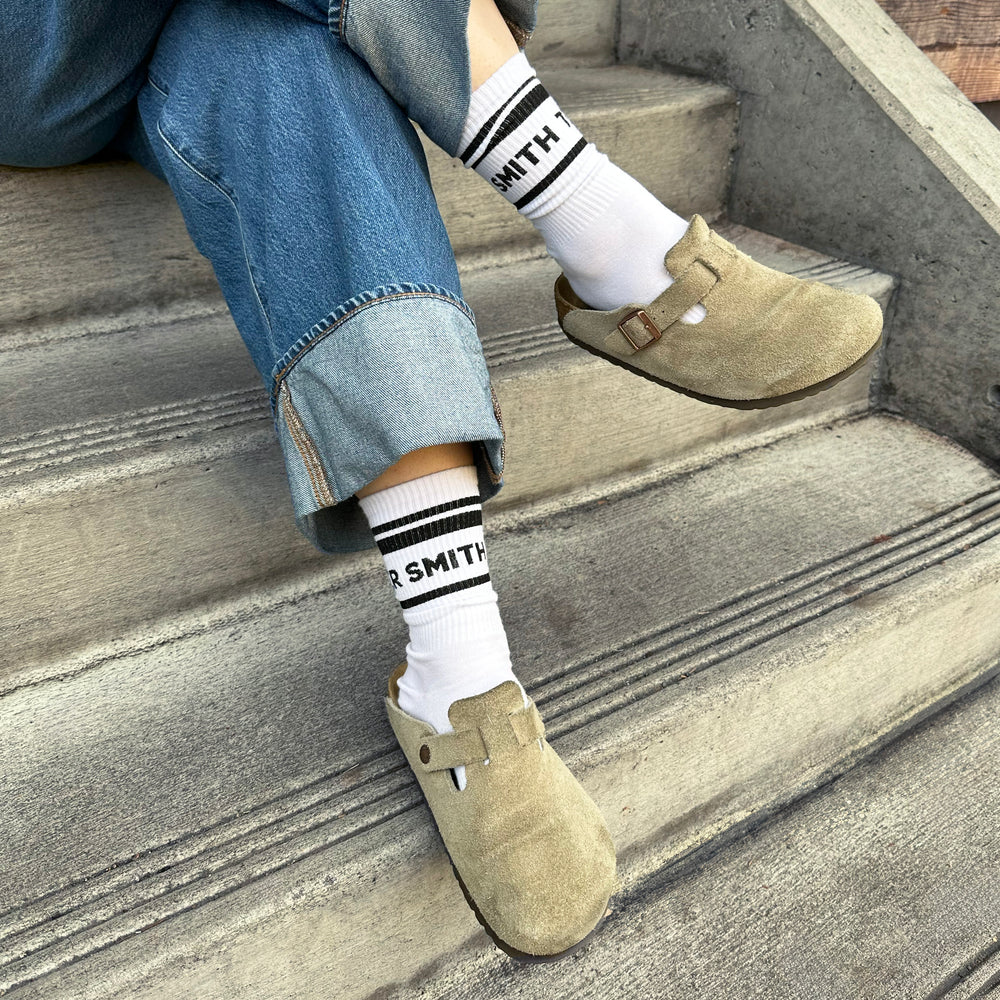 Person sitting on stairs wearing white crew socks with Smith Teamaker written around the ankle in black block letters.