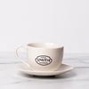 Porcelain teacup and saucer with black Smith logo shield on the front of teacup..