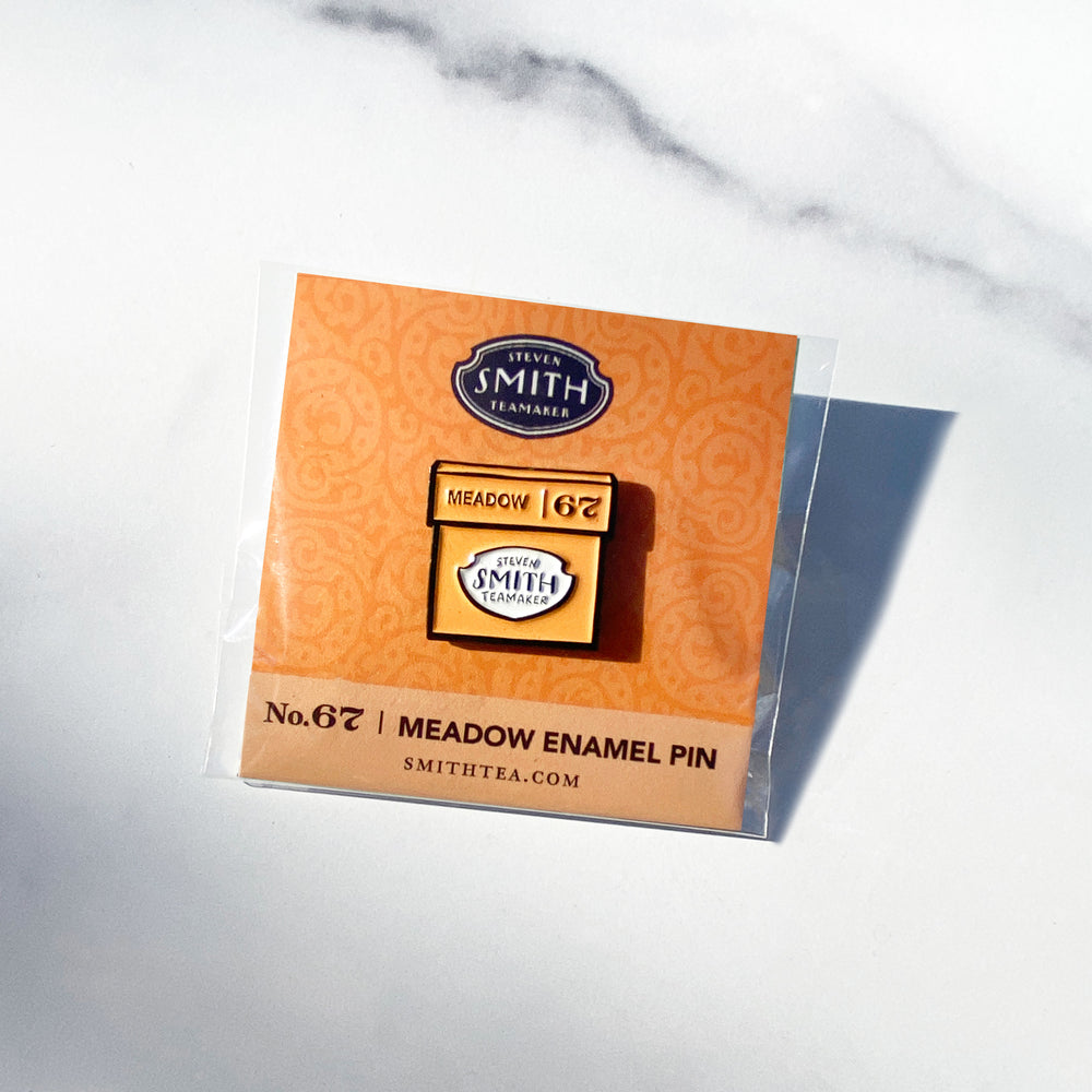 Enamel pin of an orange box of Meadow herbal infusion on an orange card with text "Meadow Enamel Pin".