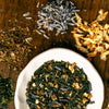Small bowl of loose leaf Tokyo Twilight green tea blend with individual ingredients staged around the bowl to show lavender, lemon peel, green rooibos and sencha green tea.