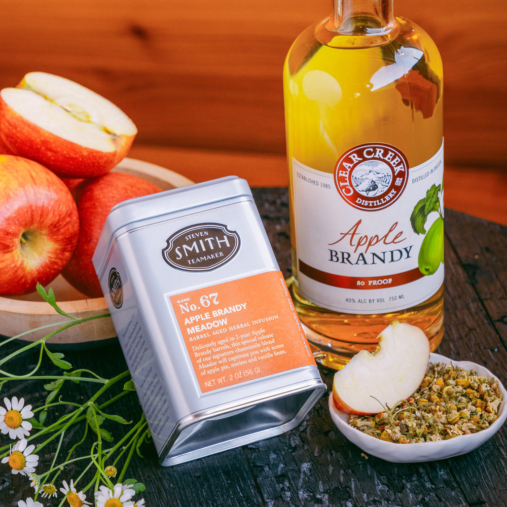 Grey tin of Apple Brandy Meadow and bottle of Apple Brandy next to a bowl of loose Meadow tea, a bowl of apples and fresh chamomile flowers.