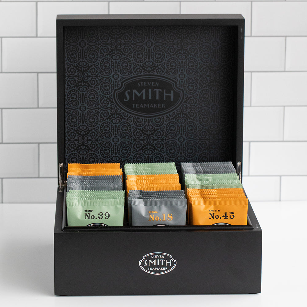 Wooden black box on kitchen counter with lid opened to showcase green, black and orange sachets of tea.