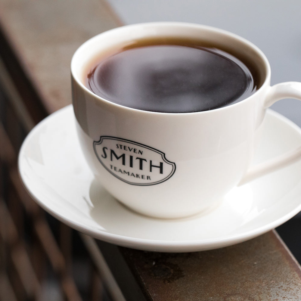 Porcelain teacup and saucer with black Smith logo shield on the front of teacup filled with black tea.