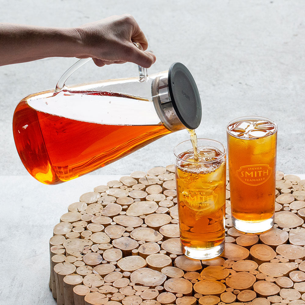 Person pouring glass pitcher with black lid filled with light brown iced tea.