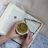 Person holding cup of Lullaby tea with sachet in the cup reading a book.