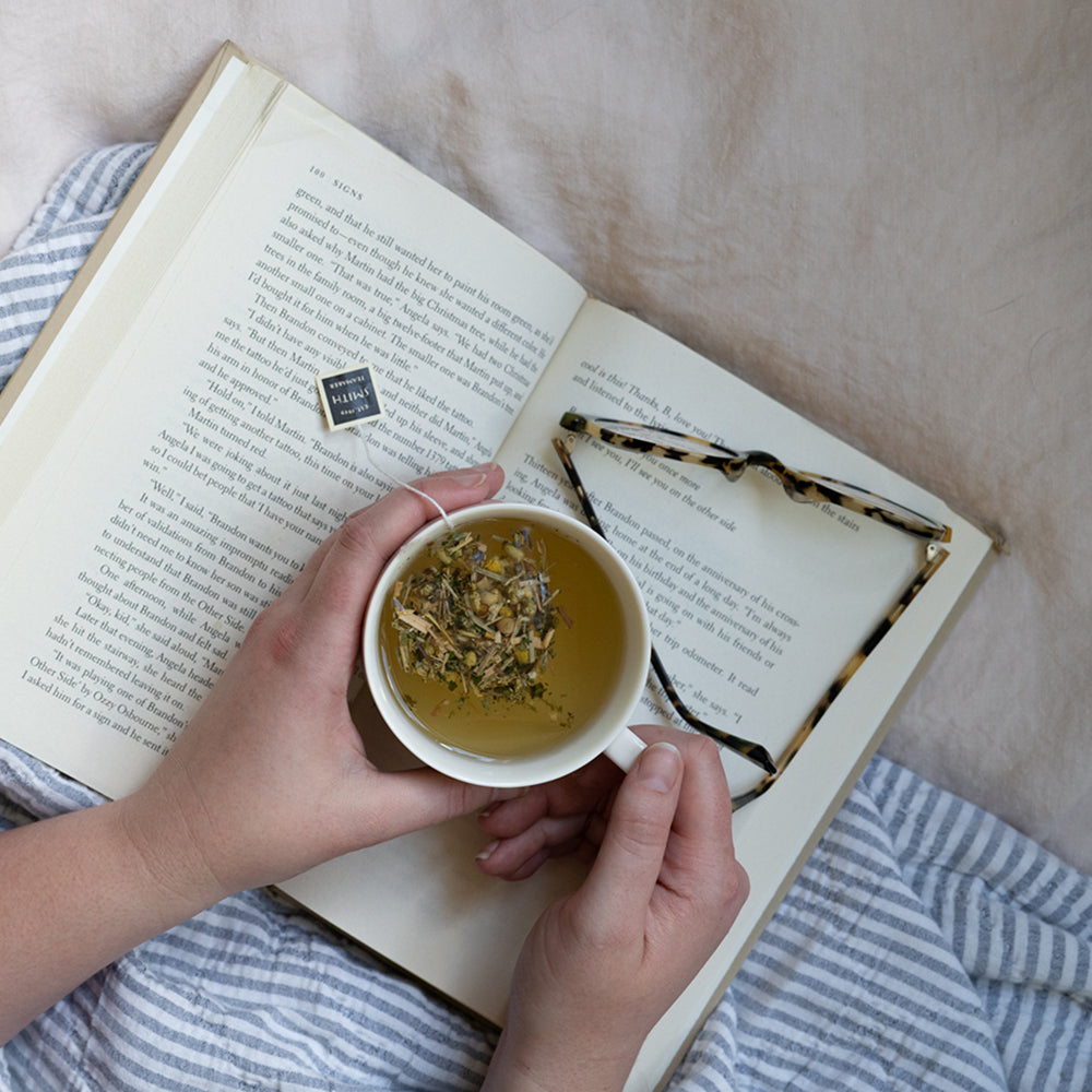 Person holding cup of Lullaby tea with sachet in the cup reading a book.