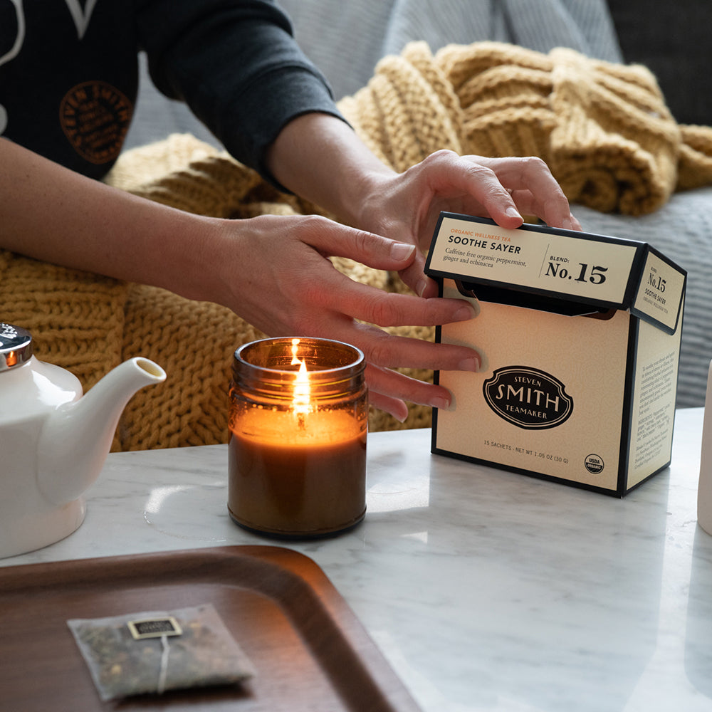 Person on a couch with yellow blanket opening cream carton of Soothe Sayer tea.