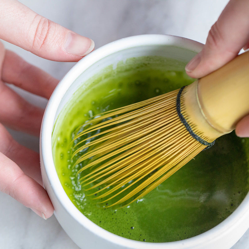 Hand holding matcha whisk stirring green matcha liquid.